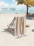 Beige towel draped over a beach chair on a sandy beach with palm trees and ocean in the background, featuring 'Lane Linen' branding.