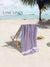 Purple towel draped over a beach chair on a sandy beach with palm trees and clear blue sky.