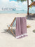 Purple towel draped over a beach chair on a sandy beach with palm trees and ocean in the background, featuring 'Lane Linen' branding.