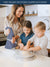 A Woman Holding Blue Color White Striped Kitchen Towel in hand and two children in a kitchen preparing food together, that indicates the heart of every happy kitchen is through Lane Linen Kitchen Towels