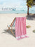 Pink towel draped over a beach chair on a sandy beach with palm trees and clear blue sky.