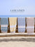 Four beach towels hanging on a wooden rack with a clear blue sky and ocean in the background, featuring the 'Lane Linen' brand.