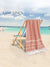 Pink towel with white stripes draped over a wooden chair on a beach, with 'Lane Linen' branding.