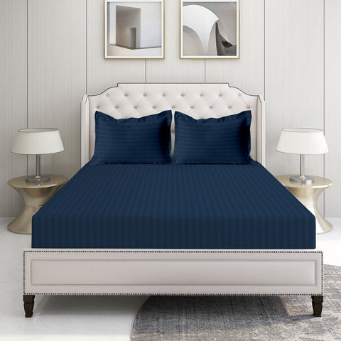 A denim blue bed sheet set on a bed in a room with a white headboard, two lamps on the nightstands, and framed pictures on the wall.
