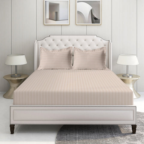 A set of Sephia Rose Color striped bed sheets displayed on a bed in a room with a white headboard and a nightstand with a lamp on the side.