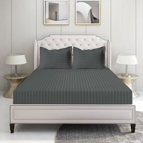 A king-sized fitted sheet set in charcoal color with a satin edge, displayed on a bed in a room with two framed pictures on the wall and a nightstand with a lamp.