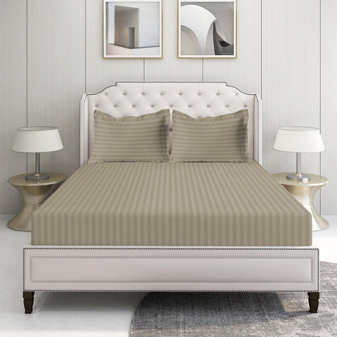 A taupe-colored, 100% organic cotton king fitted sheet set with two pillowcases, displayed on a bed in a room with a white headboard and a nightstand with a lamp on each side.
