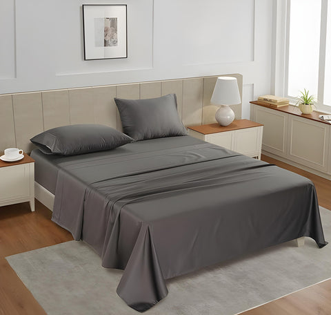 A charcoal gray king flat satin bedsheet set on a bed in a room with a tan headboard and a nightstand with a lamp and a plant beside the bed.