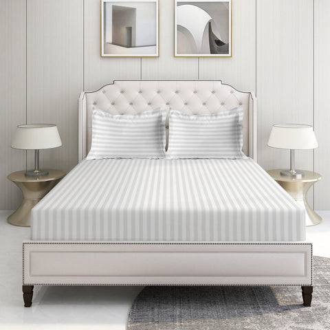 A set of White Color striped bed sheets displayed on a bed in a room with a white headboard and a nightstand with a lamp on the side.