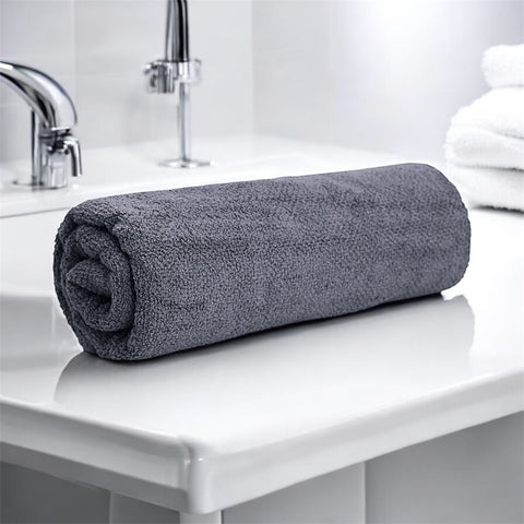 A rolled up gray bath towel placed on a white surface, likely a bathroom counter, with a faucet and partial view of a shower curtain in the background.