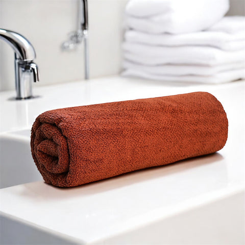 A rolled-up brown color cotton bath towel placed on a bathroom counter, with a tap and towels in the background