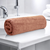 A rolled-up peach color cotton bath towel placed on a bathroom counter, with a tap and towels in the background