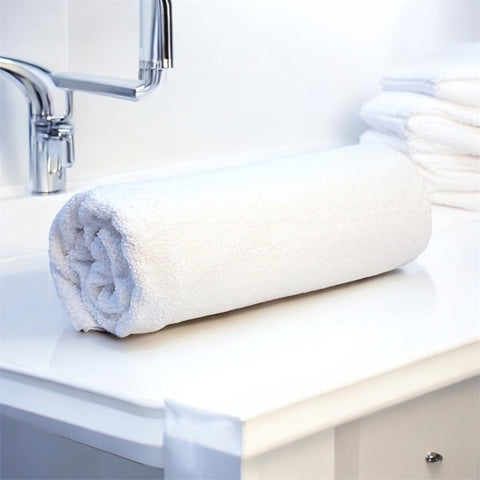 A rolled-up white cotton bath towel placed on a bathroom counter, with a tap and towels in the background.