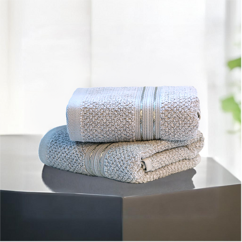 Two folded hand towels on a flat surface, with a textured silver color, placed against a backdrop of a bright window and plants.