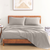 A gray cotton bed sheet set on a bed with a wooden headboard, accompanied by pillowcases. The room has a minimalistic design with a framed picture on the wall and bedside tables.